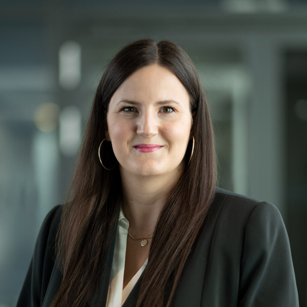 Zu sehen ist eine Frau mit langen dunklen Haaren die sie offen trägt. Sie reichen ihr bis zur Brust. Sie hat trägt große Creolen Ohrringe, eine dezente Kette, einen schwarzen Blazer und eine weiße Bluse. Es handelt sich um Prof. Dr. Carolin Strassmann die im Transferhub arbeitet. 